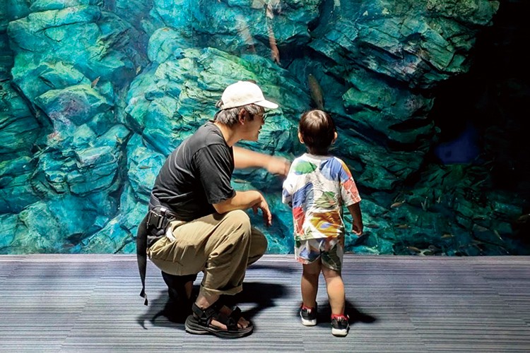 写真：水族館の水槽の前で、小さな子どもに話しかける帽子をかぶった男性。後ろ姿で、2人は水中の生き物を見ているようす。