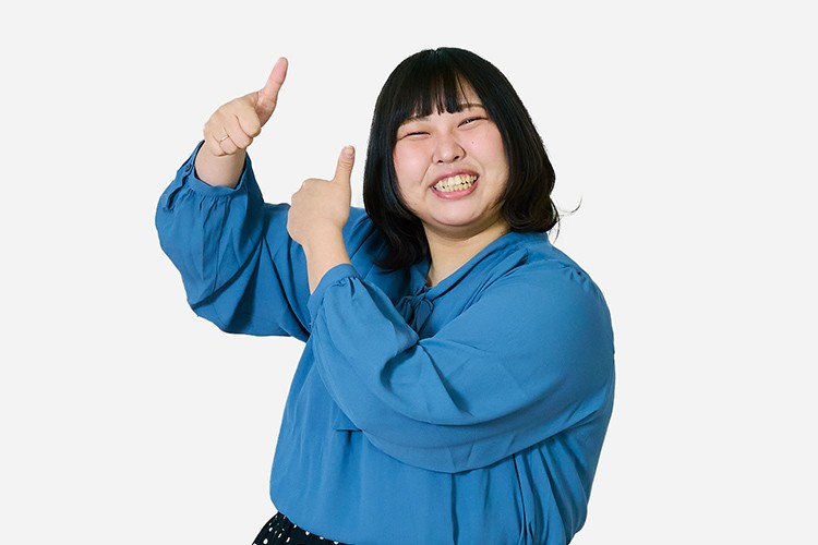 写真：青いブラウスを着た笑顔の女性。両手で親指を立ててポーズをとっている。