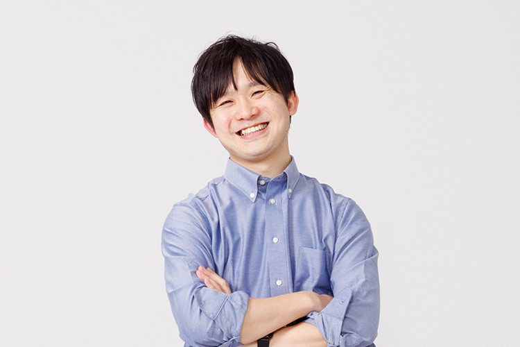 写真：青いシャツを着た男性が腕を組んで笑顔を見せている。