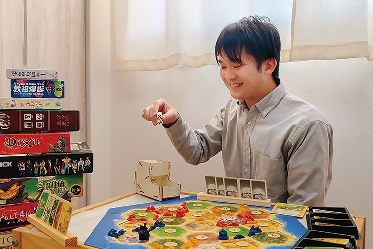 写真：ボードゲーム『カタン』を楽しむ男性。サイコロを振って笑顔を見せている。周囲にはたくさんのボードゲームが積まれている。