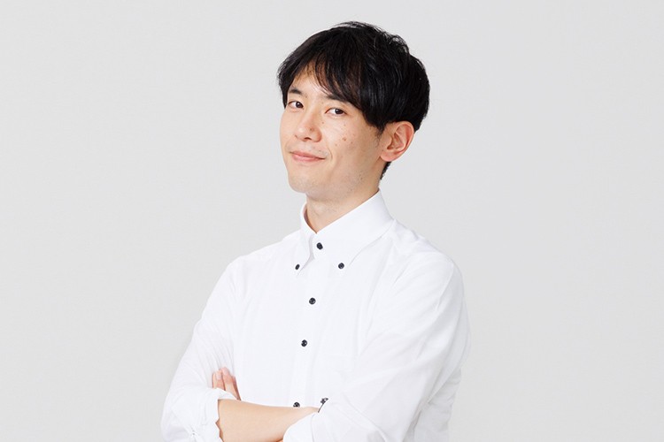 写真：白いシャツを着た男性が腕を組んで微笑んでいる。