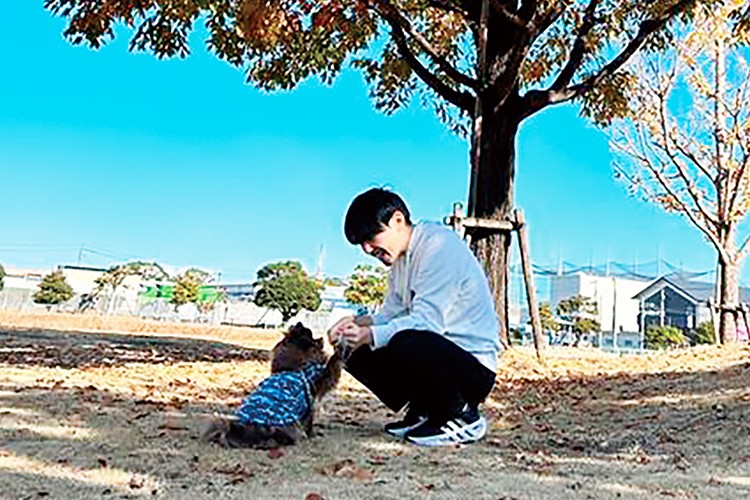 写真：秋の公園で犬と向き合う男性。しゃがんで犬と握手しているようす。背景には青空と紅葉した木々が広がる。
