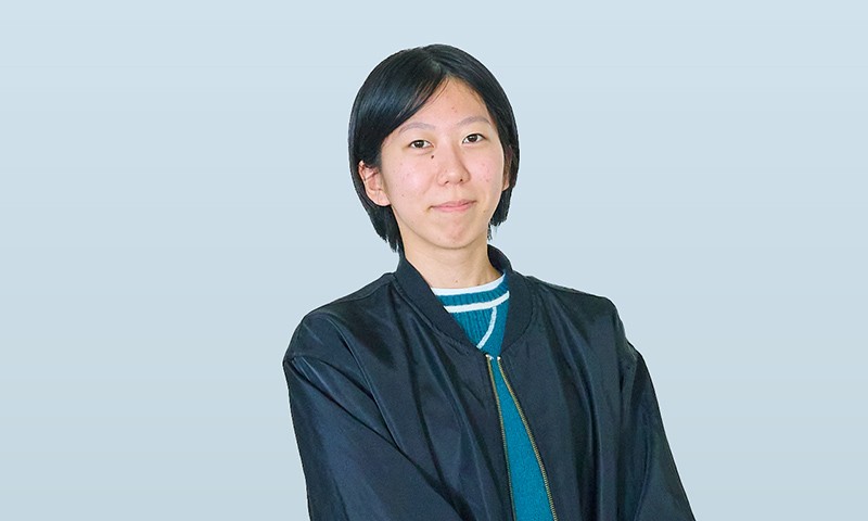 写真：黒いジャケットを着た女性学生。淡いブルーの背景の前で、静かにカメラを見つめている。
