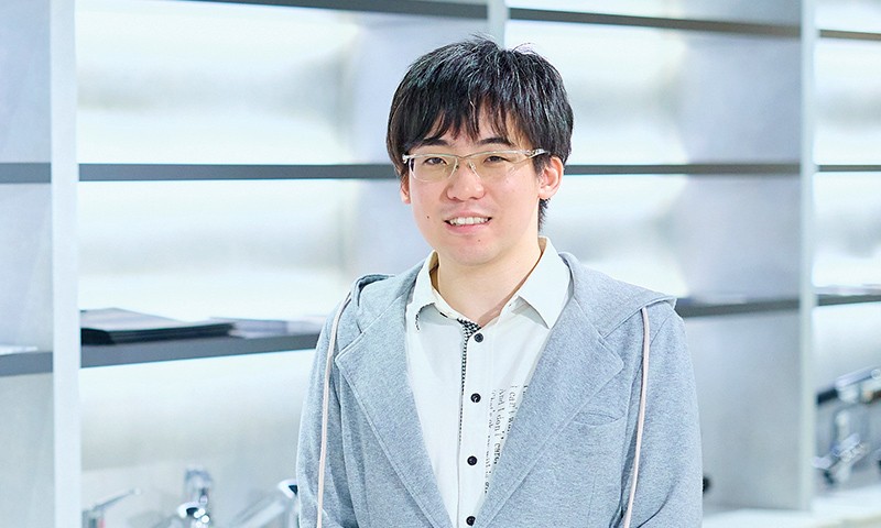 写真：白いシャツにグレーのパーカーを重ねた男性。メガネをかけ、落ち着いた雰囲気で笑顔を見せている。
