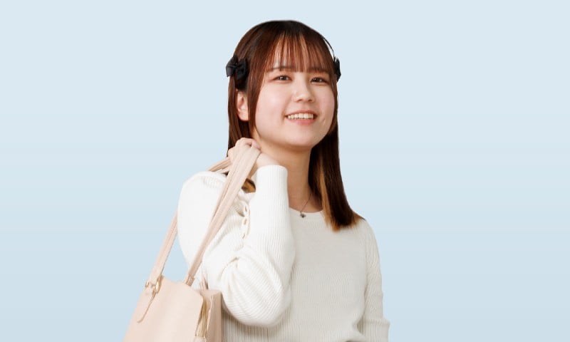写真：白いトップスを着た女性学生。ベージュのバッグを肩にかけて、微笑んでいる。背景は淡いブルー。
