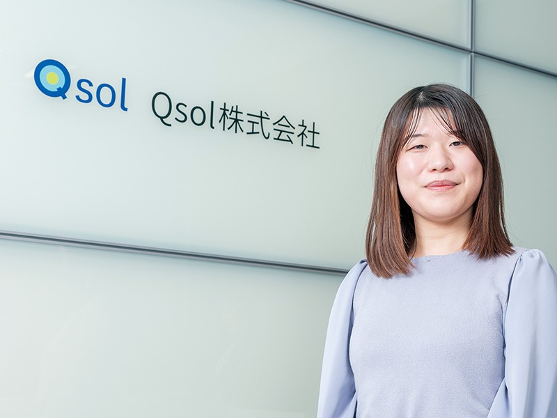 写真：薄灰色の服を着た笑顔の女性。後ろの壁にQsol株式会社のロゴマークと社名が掲げられている。