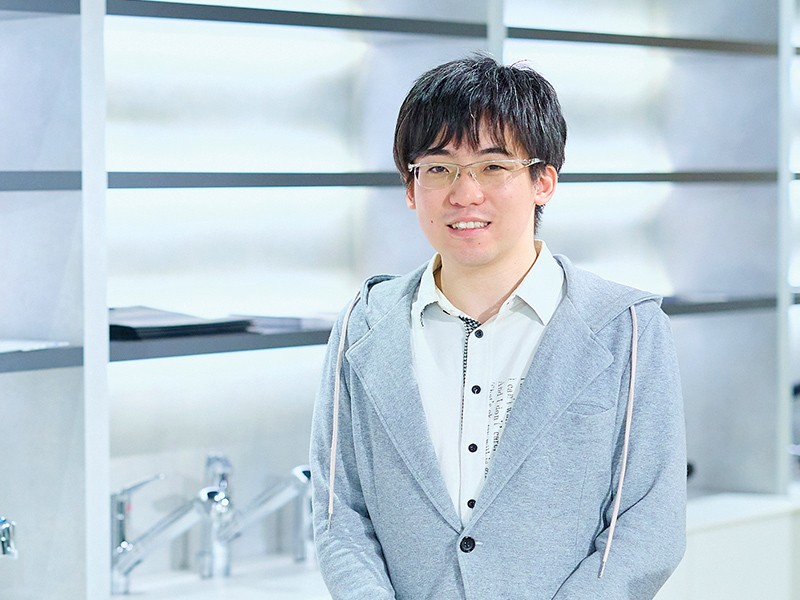 写真：白いシャツにグレーのパーカーを重ねた男性。メガネをかけ、落ち着いた雰囲気で笑顔を見せている。