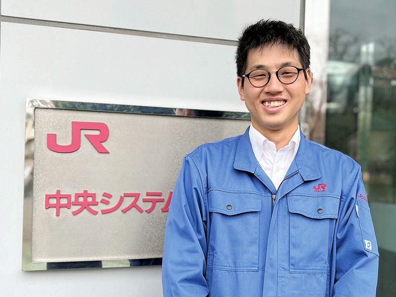写真：青い制服を着た男性。JR中央システムの看板の前で、満面の笑顔を浮かべて立っている。