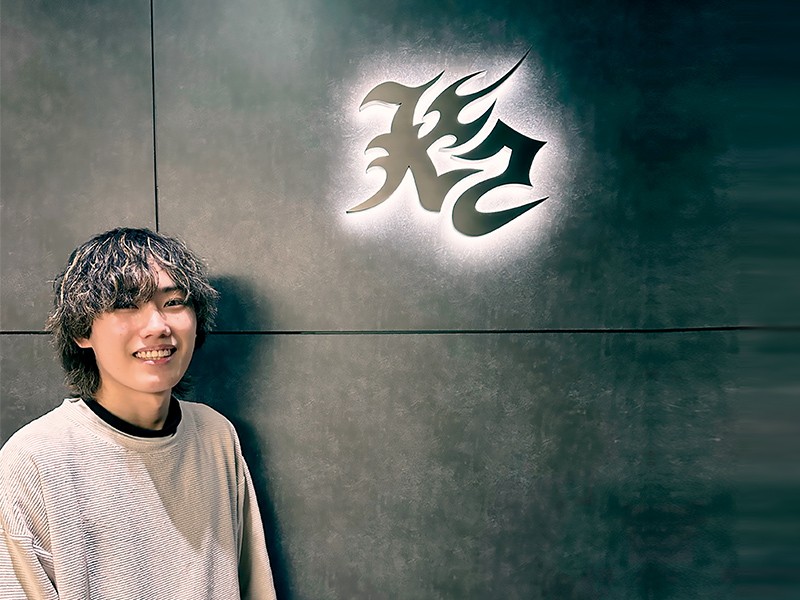 写真：ライトベージュのトップスを着た男性。壁に浮かび上がる株式会社ケーツーのロゴの前で、柔らかい笑顔を見せている。