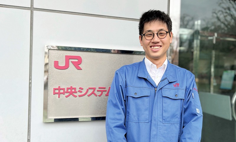 写真：青い制服を着た男性。JR中央システムの看板の前で、満面の笑顔を浮かべて立っている。
