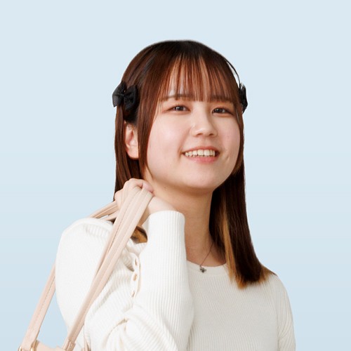 写真：白いトップスを着た女性学生。ベージュのバッグを肩にかけて、微笑んでいる。背景は淡いブルー。