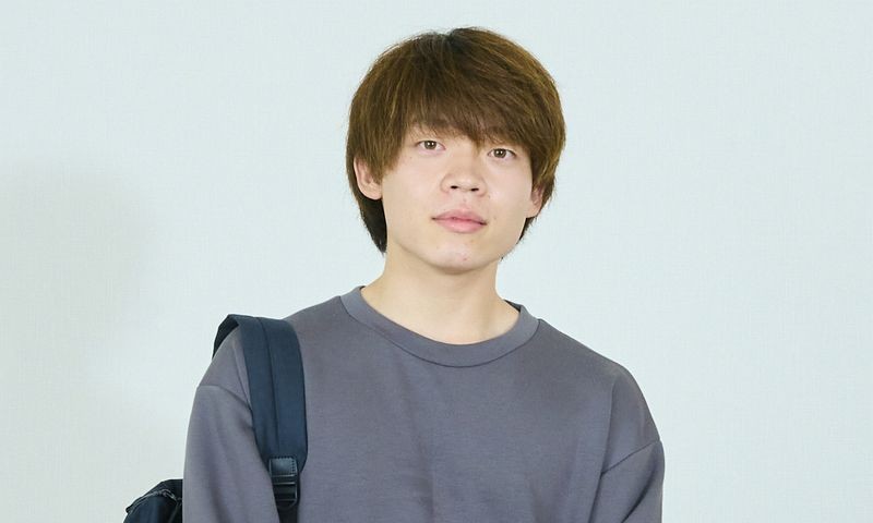 写真：グレーのスウェットを着た男子学生。スマートフォンで電話をかけている。